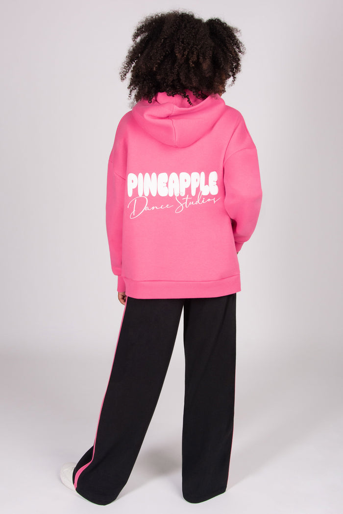 Girl wearing a pink oversized hoodie with 'Pineapple Dance Studios' print on the back, standing against a white background.