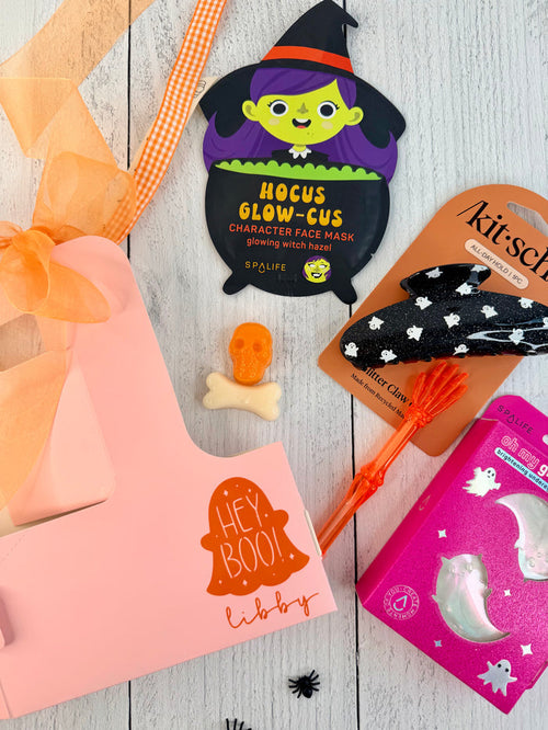 A styled flat-lay of Halloween-themed beauty products on a white wood background, including a pink gift box with orange ribbon, a witch face mask packet, a black star-patterned headband on a card, an orange hairbrush, and a pink ghost box.