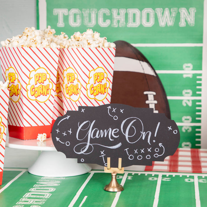 A football-themed snack table featuring a Black Frame Table Accent standing in a place card holder, with "Game On!" hand-written in white script along with accents of Xs and Os with arrows in the style of football strategy maps.