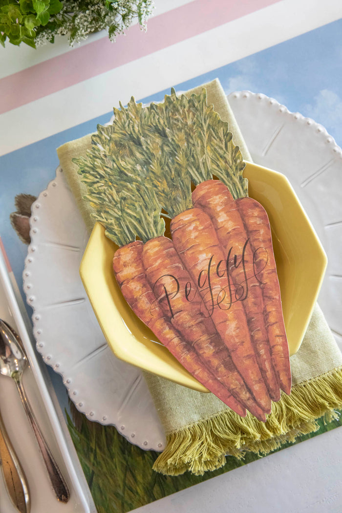A Carrots Table Accent labeled "Peggy" resting on the plate of a springtime place setting, from above.