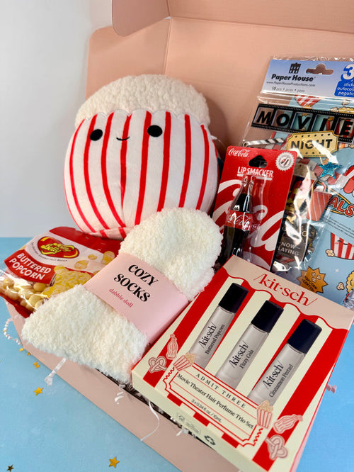A curated gift box filled with themed items, including a plush character in red and white stripes, a pink sock, a KitSch perfume set, a bag of Pop-Tart, and a bag of Kicker's Butter Popcorn, all arranged in a pink box.