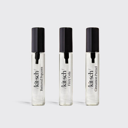 Three clear glass roll-on perfume bottles with black caps, labeled Kit's Cheddar Buttered Popcorn, Kit's Cheddar Fizzy Cola, and Kit's Cheddar Cinnamon Pretzel, arranged on a light gray background.