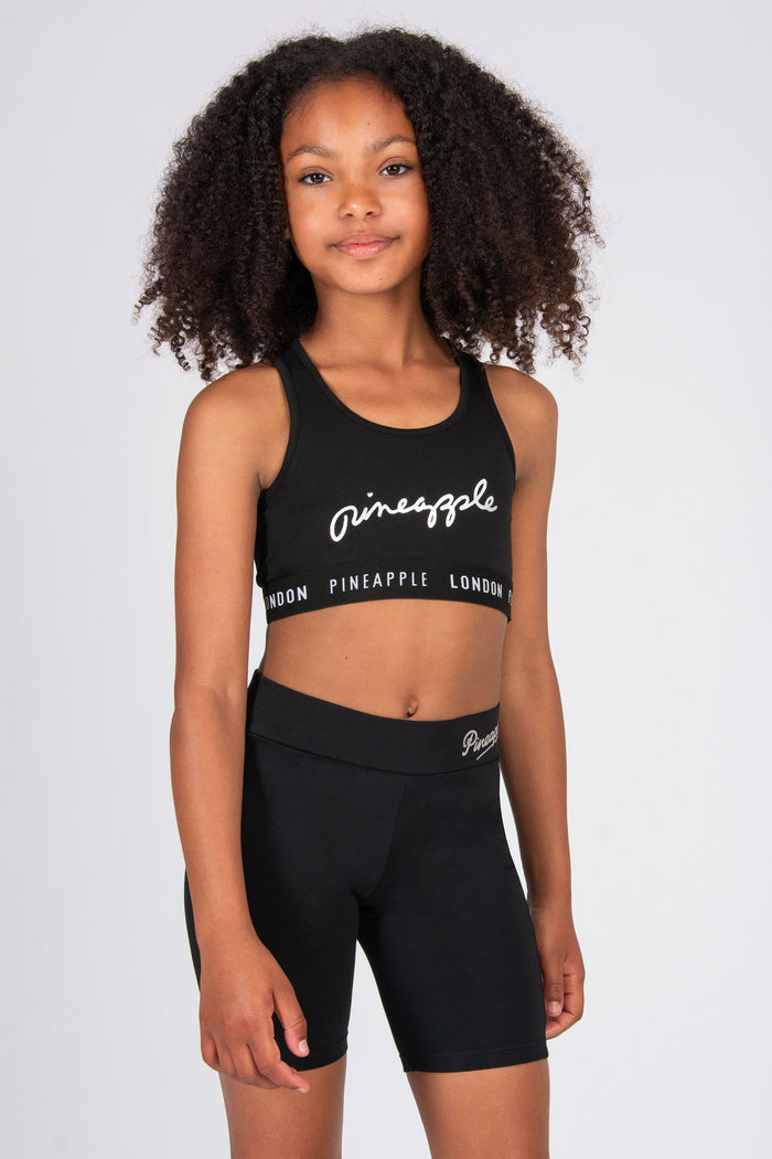 Young girl wearing a black sports bra and cycling shorts with 'Pineapple London' branding on a white background