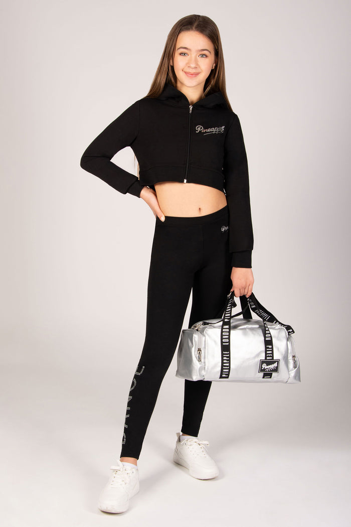 Girl in black athletic outfit holding a silver duffel bag on a white background