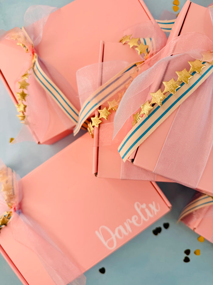 Personalized pink gift boxes with tulle ribbon, gold star garlands, and blue stripes, arranged on a light blue background. One box is printed with the name 'Darelix'.