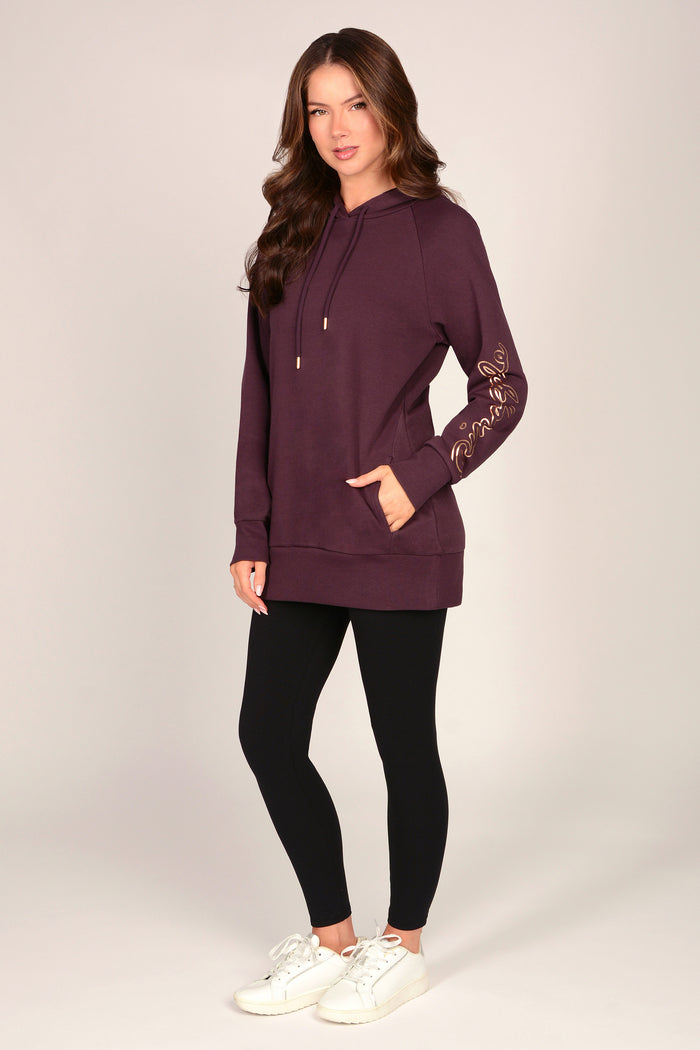 Woman wearing a maroon brown hoodie with pockets and black leggings against a grey background