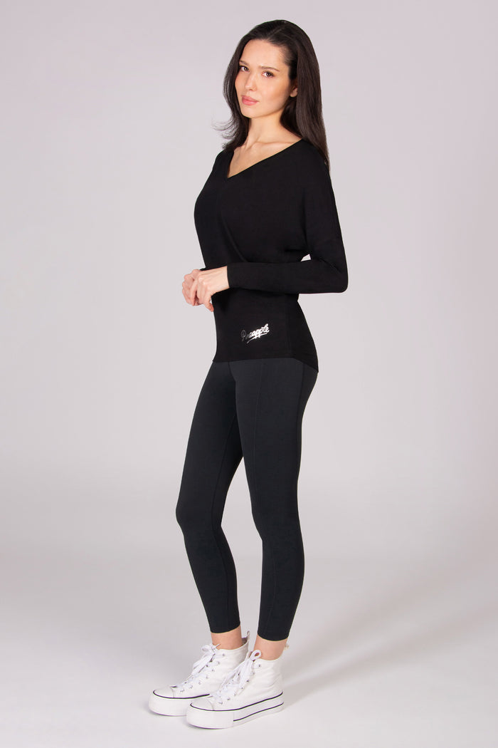 Woman wearing a long sleeve black top with a v neckline and black leggings paired with white sneakers on a light grey background
