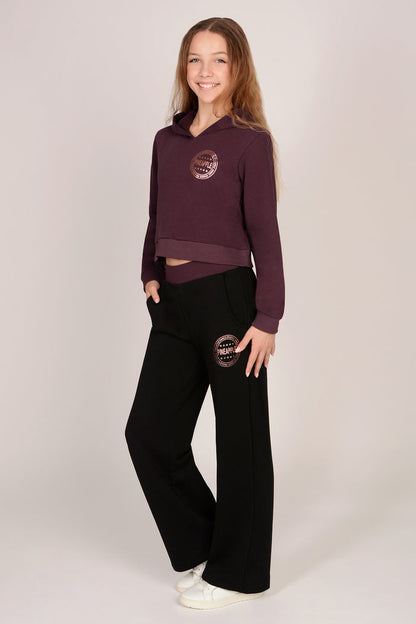 Teenage girl wearing a maroon brown hoodie with matching black wide leg trousers against a plain background