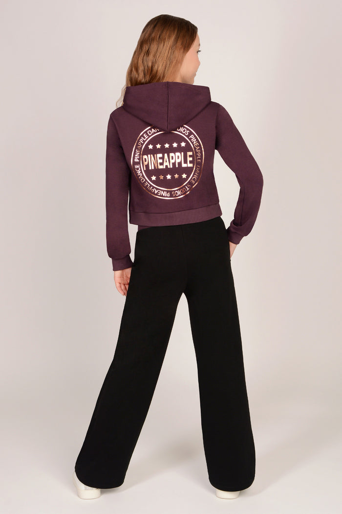 Teenage girl wearing a maroon brown hoodie with matching black wide leg trousers against a plain background
