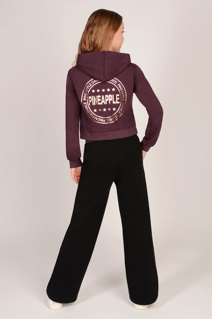 Teenage girl wearing a maroon brown hoodie with matching black wide leg trousers against a plain background