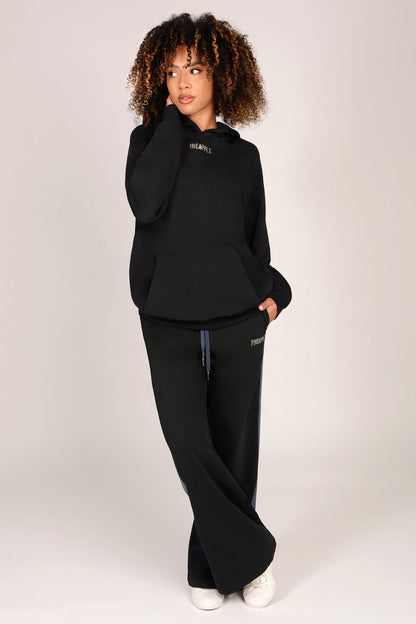 Woman wearing a black tracksuit consisting of a kangaroo pocket hoodie and matching wide leg joggers on a white background