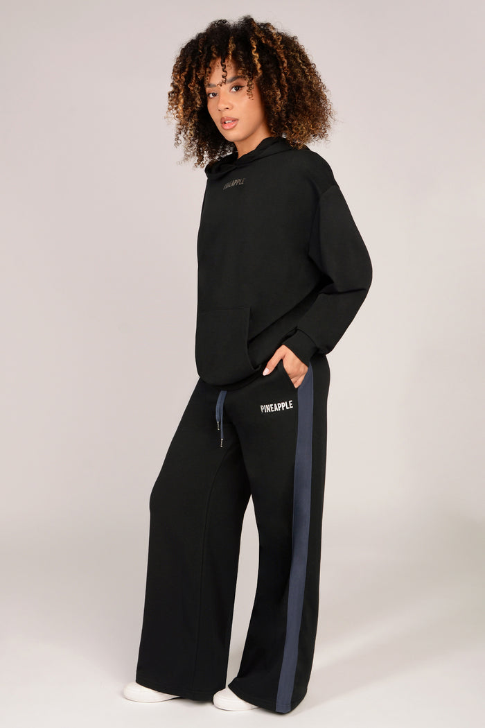 Woman wearing a black oversized hoodie with kangaroo pockets with matching wide leg black trousers with dark grey paneling running down the sides against a white background.