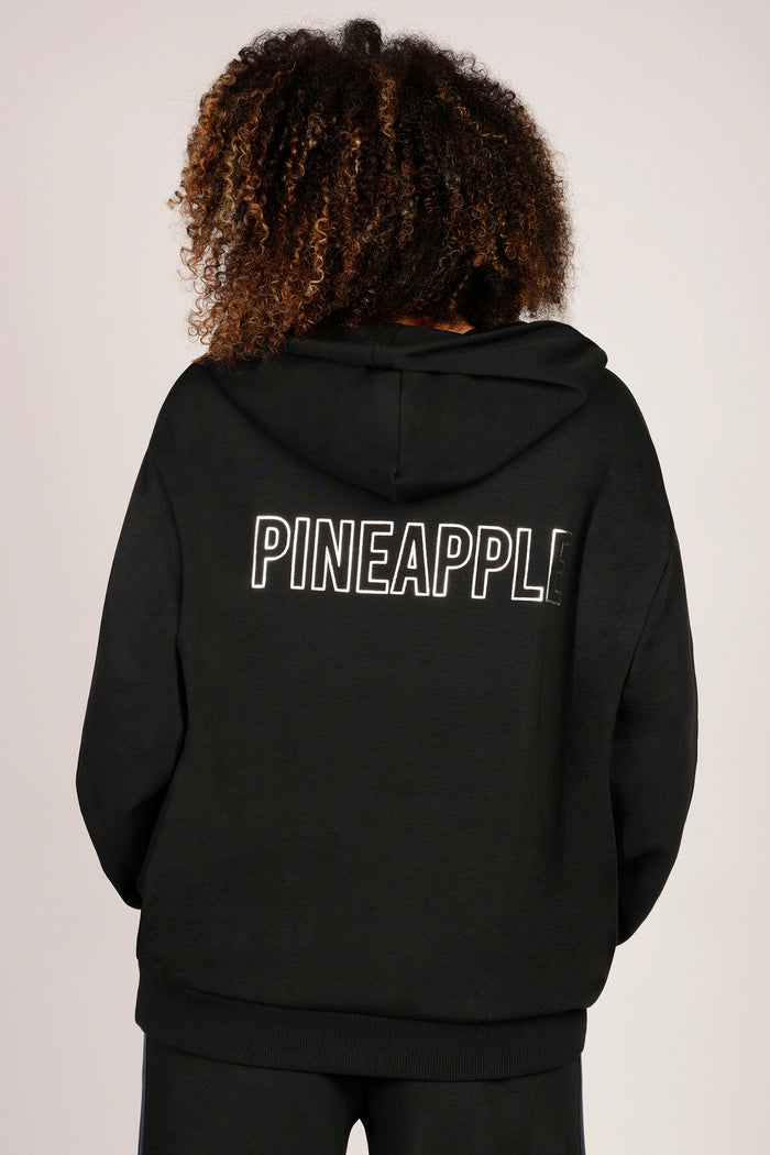 Woman wearing a black boxy hoodie with 'PINEAPPLE' printed on the back against a plain background.