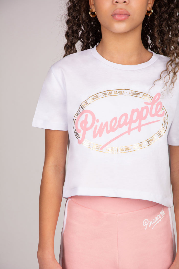 Young girl wearing a white t-shirt with 'Pineapple Dance' print and light pink shorts on a light grey background