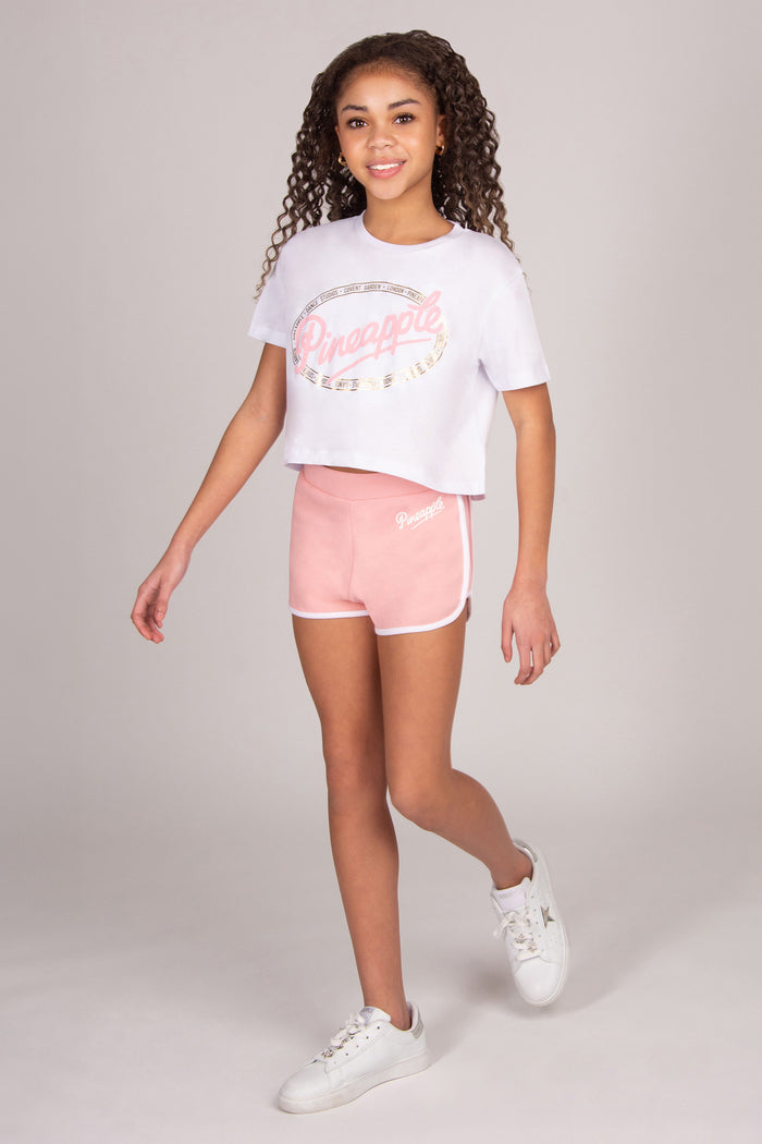 Young girl wearing a white t-shirt with 'Dance' print and light pink shorts on a light grey background