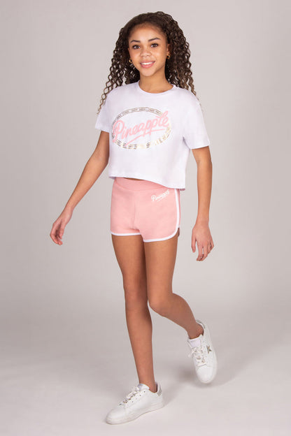 Young girl wearing a white t-shirt with 'Dance' print and light pink shorts on a light grey background