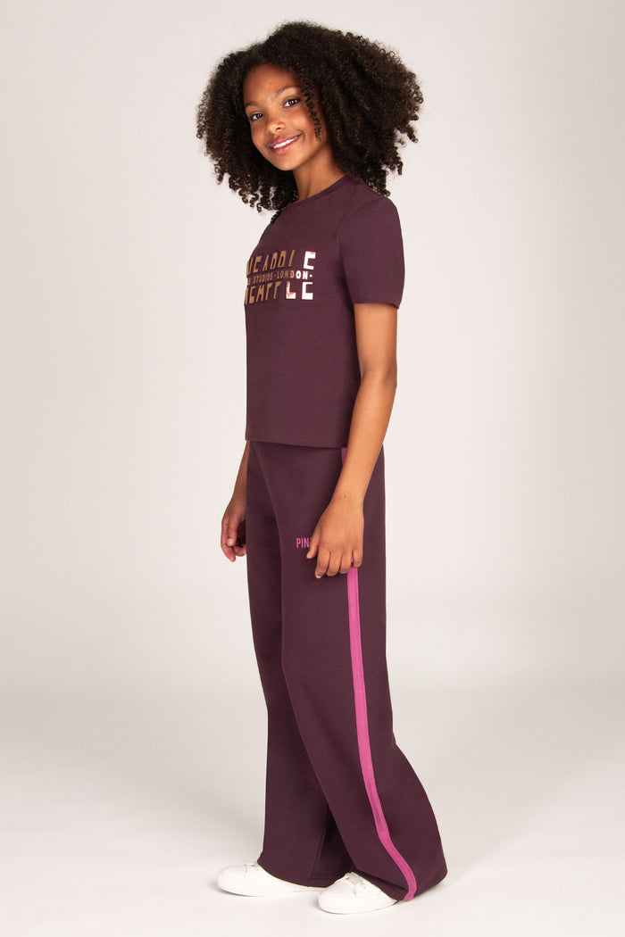 Girl wearing maroon brown trousers with pink side panel and matching t-shirt on a plain background