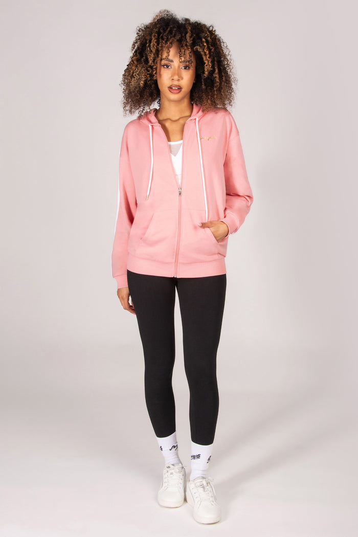 Woman wearing a light pink hoodie with zip and pockets, black leggings, and white sneakers on a light grey background
