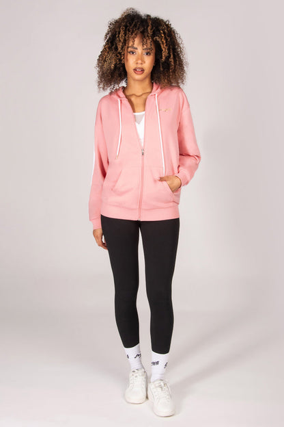 Woman wearing a light pink hoodie with zip and pockets, black leggings, and white sneakers on a light grey background