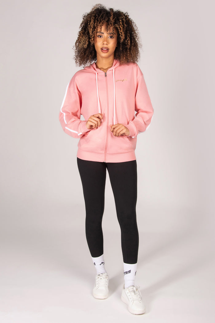 Woman wearing a light pink zip hoodie, black leggings, and white sneakers on a light grey background