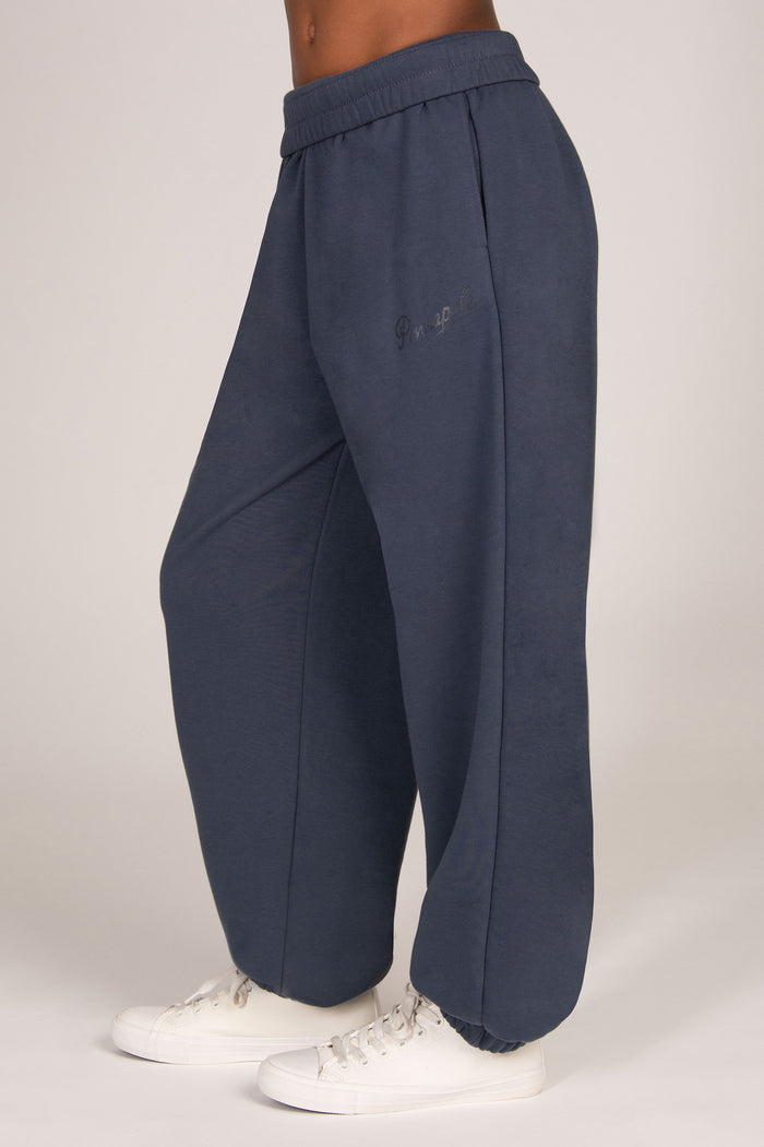 A person wearing loose, wide-leg sweatpants in a muted blue-grey color, paired with white sneakers, shown from the waist down against a plain light background.