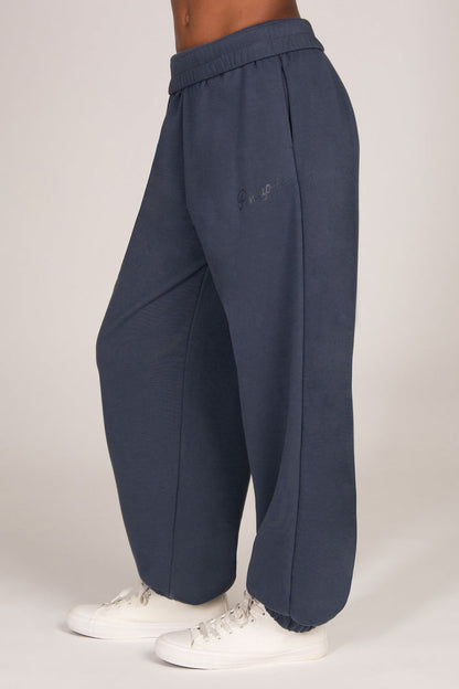 A person wearing loose, wide-leg sweatpants in a muted blue-grey color, paired with white sneakers, shown from the waist down against a plain light background.