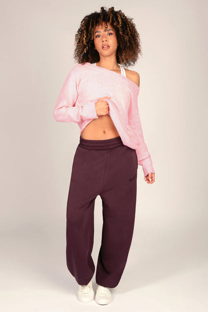 A woman modelling a pair of oversized dark aubergine joggers with a light pink knitted sweater and white trainers, standing against a light grey studio background.