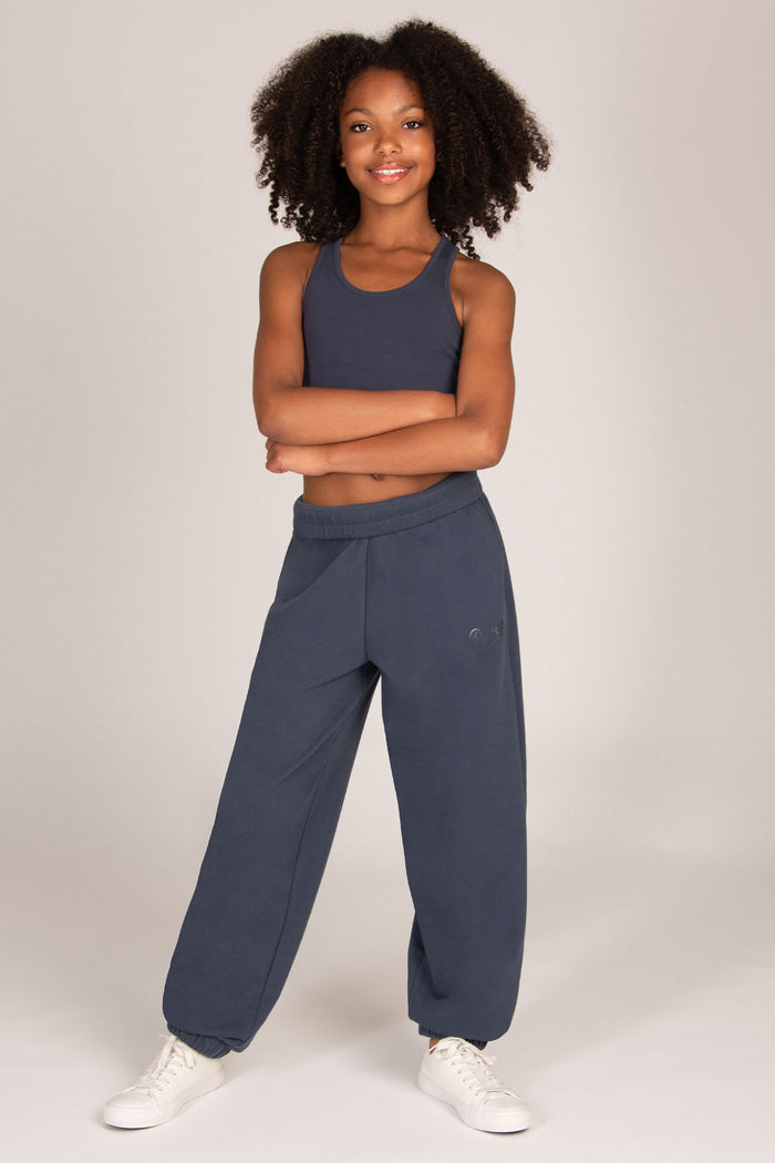 A young girl stands against a light grey studio background wearing a matching charcoal grey activewear set consisting of a cropped tank top and wide-leg oversized balloon sweatpants, paired with white sneakers.