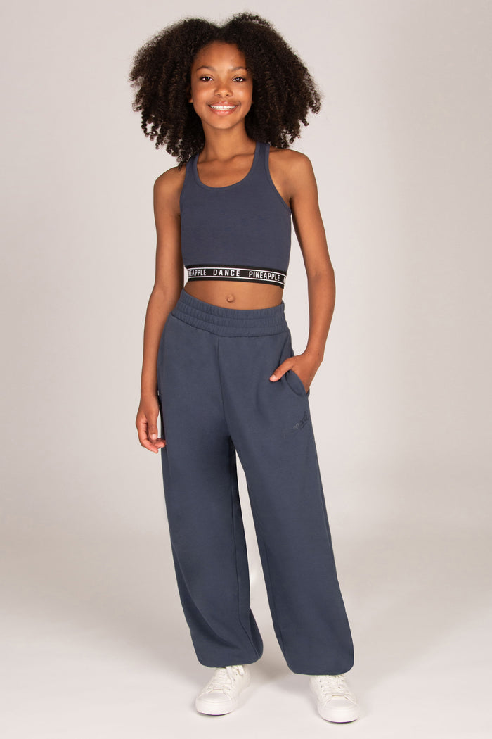 A young girl stands against a light grey studio background wearing a matching charcoal grey activewear set consisting of a cropped tank top and wide-leg oversized balloon sweatpants, paired with white sneakers.