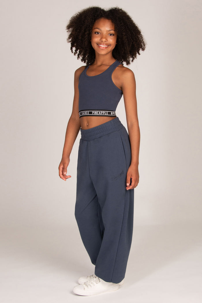 A young girl stands against a light grey studio background wearing a matching charcoal grey activewear set consisting of a cropped tank top and wide-leg oversized balloon sweatpants, paired with white sneakers.