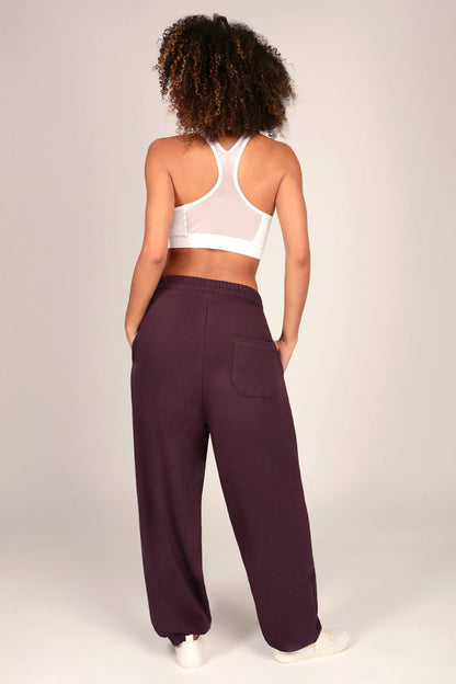 A woman modeling a pair of dark plum sweatpants with a white sports bra and white sneakers, standing against a light grey studio background.