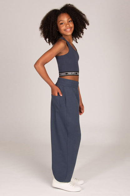 A young girl stands against a light grey studio background wearing a matching charcoal grey activewear set consisting of a cropped tank top and wide-leg oversized balloon sweatpants, paired with white sneakers.