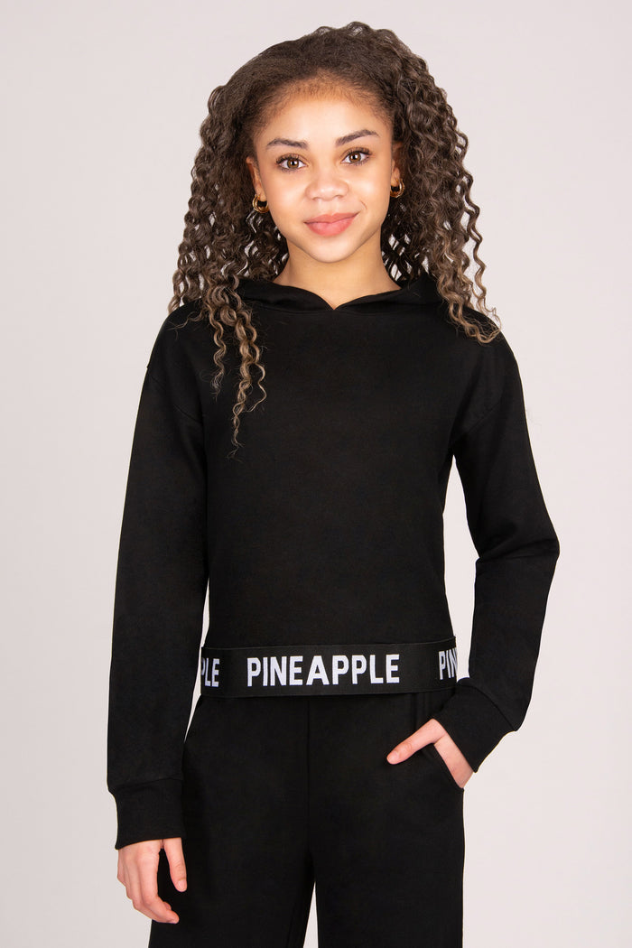 Girl wearing a black lightweight hoodie with 'Pineapple Dance' tape hem.