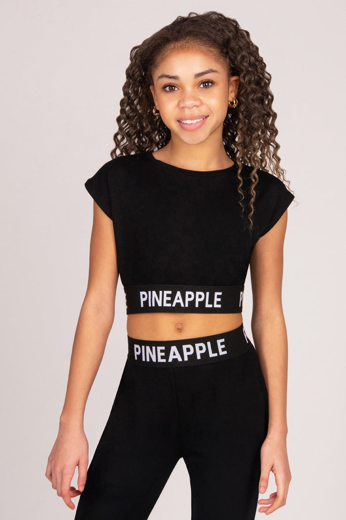 Girl wearing a black crop top and matching trousers with 'PINEAPPLE' branding on a plain background