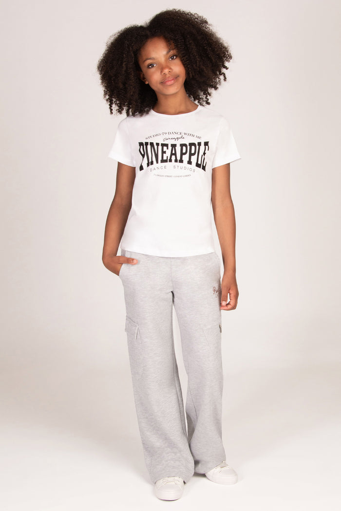 A young girl wearing a white short-sleeve graphic t-shirt with 'PINEAPPLE' printed in black across the chest, paired with light grey wide-leg sweatpants with cargo pockets and white sneakers.