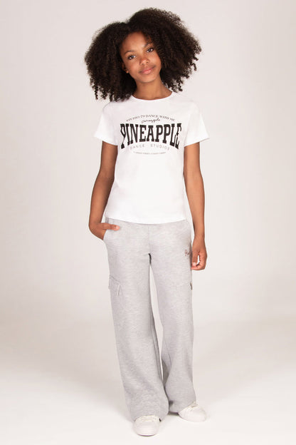 A young girl wearing a white short-sleeve graphic t-shirt with 'PINEAPPLE' printed in black across the chest, paired with light grey wide-leg sweatpants with cargo pockets and white sneakers.